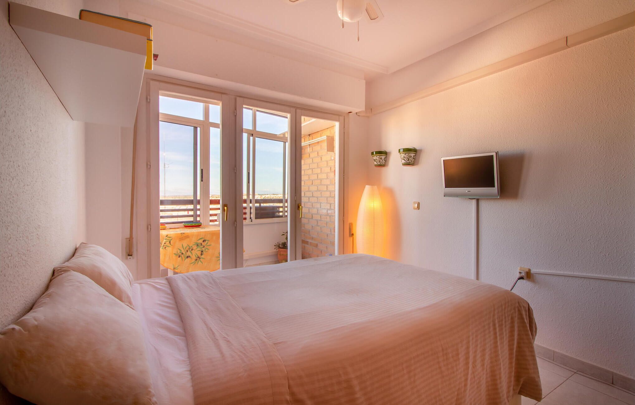 Ein Schlafzimmer mit Bett, Fernseher, einem Balkon mit Tisch und Stühlen sowie einer Tür zum Ausgang.