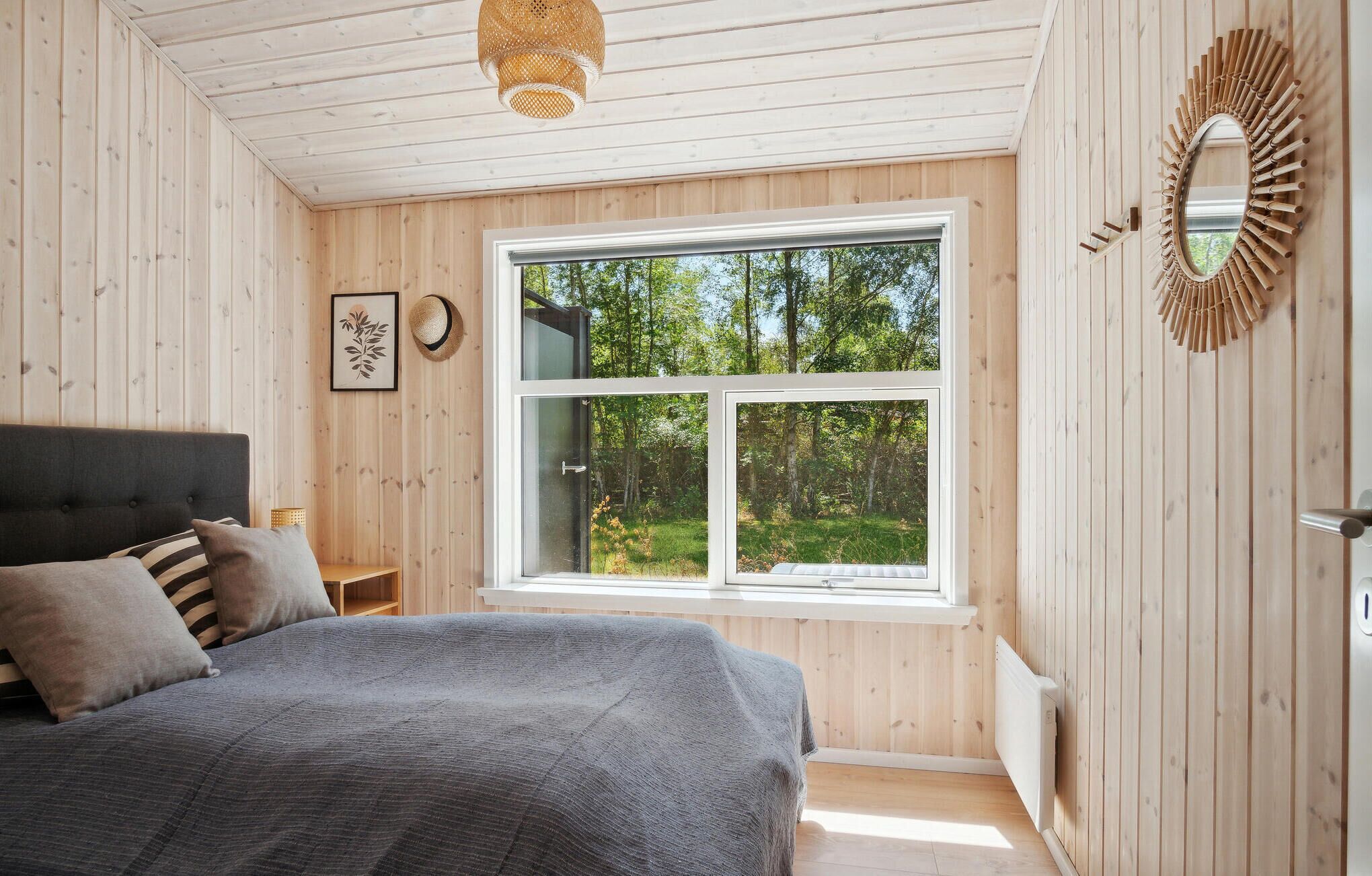 Une chambre avec une grande fenêtre, des murs en bois, un lit avec une literie grise, une table de chevet et un luminaire suspendu.