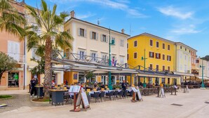 Un café en plein air, avec des tables et des chaises, entouré d’immeubles dotés de balcons et de volets.