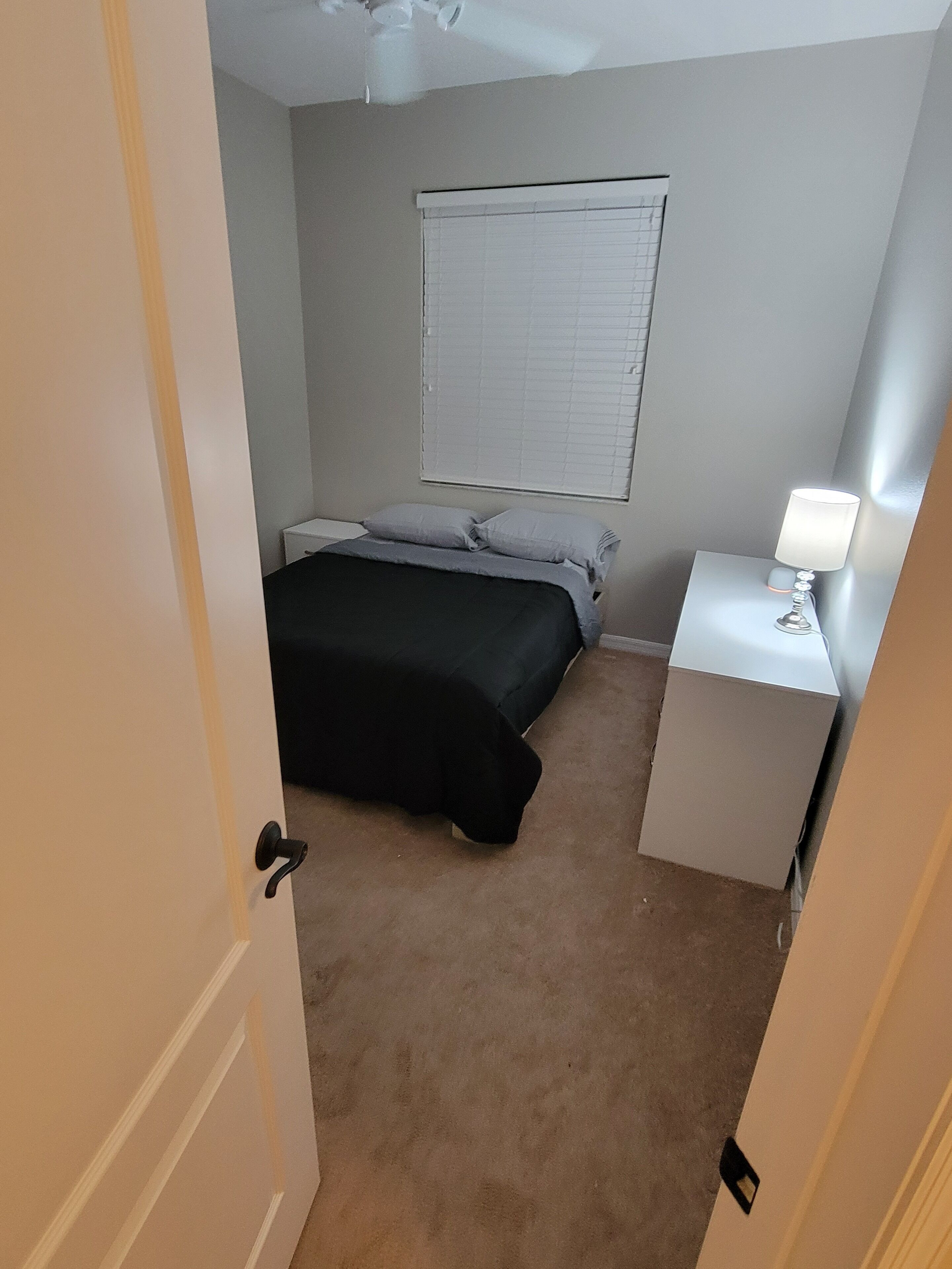 A bedroom with a bed, a bedside table with a lamp, a window with blinds, and a door.