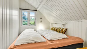 Un dormitorio con techo inclinado, una ventana con vistas a la vegetación, una cama con ropa de cama blanca y naranja, y una mesita de noche con una lámpara.
