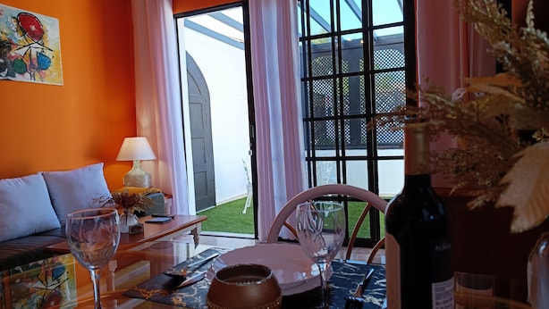 Una habitación con un sofá, una mesa con un florero y una copa de vino, y vistas a un jardín a través de ventanas grandes.