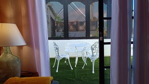 Una habitación con una mesa y sillas blancas, una lámpara y vistas a través de una ventana con celosía negra.
