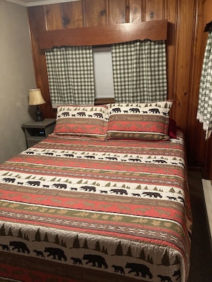 A bed with a patterned bedspread, two pillows with a bear and forest design, a nightstand with a lamp, and a window with checkered curtains. - Pymatuning Lake Walking distance to Espyville Marina! (Pymatuning Central)