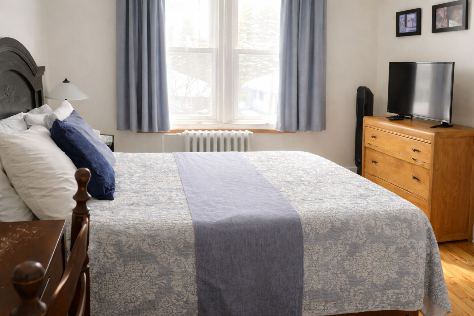 A neatly made bed with a patterned bedspread, a wooden dresser with a television, and a window with curtains.