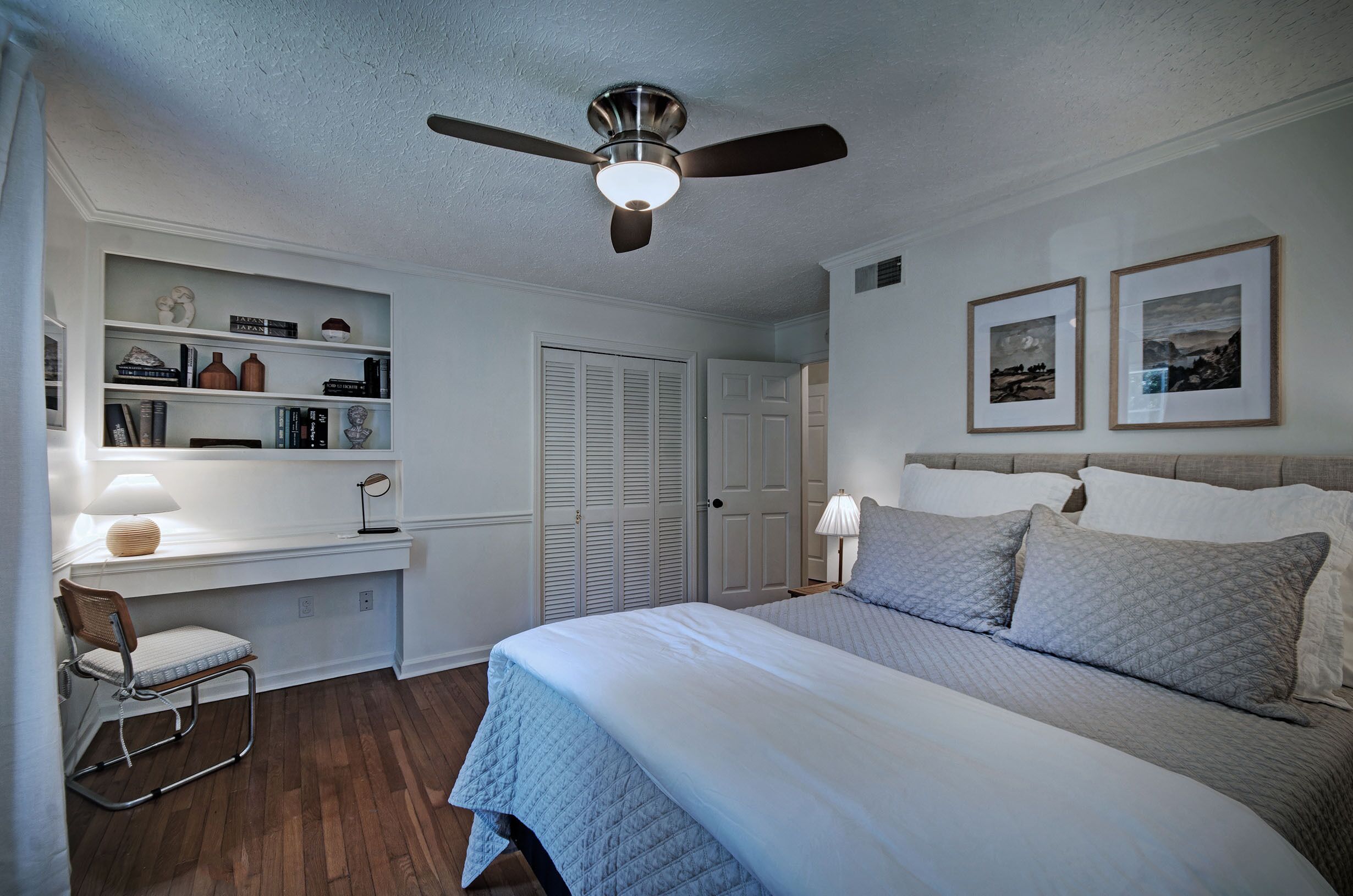 A bedroom with a bed, a desk, a chair, a ceiling fan, and a door.