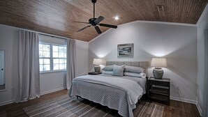 A bedroom with a wooden ceiling, a bed with a grey quilt, two bedside tables with lamps, and a window with curtains. - Serene Home in Fondren Location Near Hospitals (Jackson)