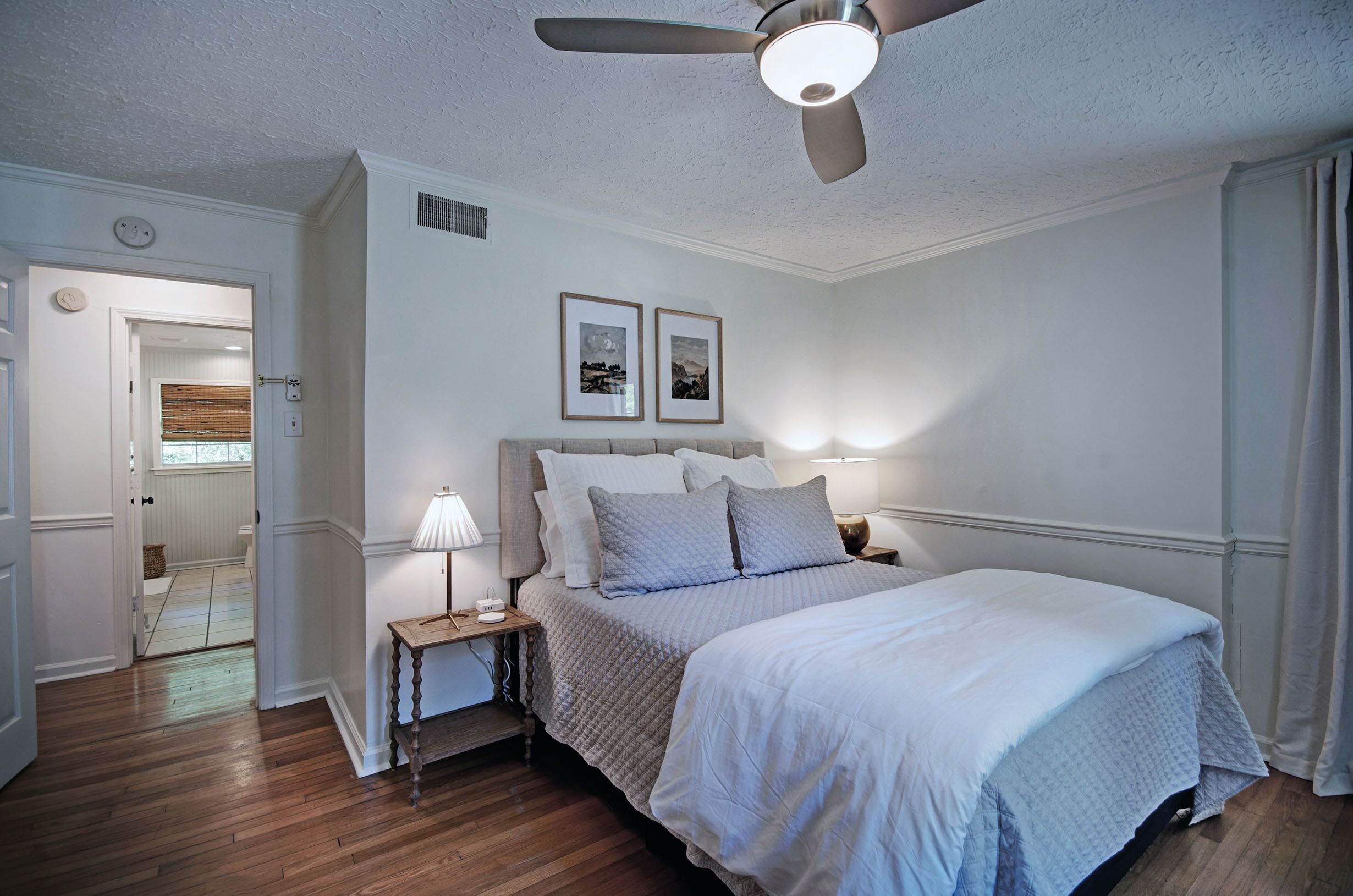 A bedroom with a bed, bedside table, lamp, ceiling fan, and framed pictures on the wall.