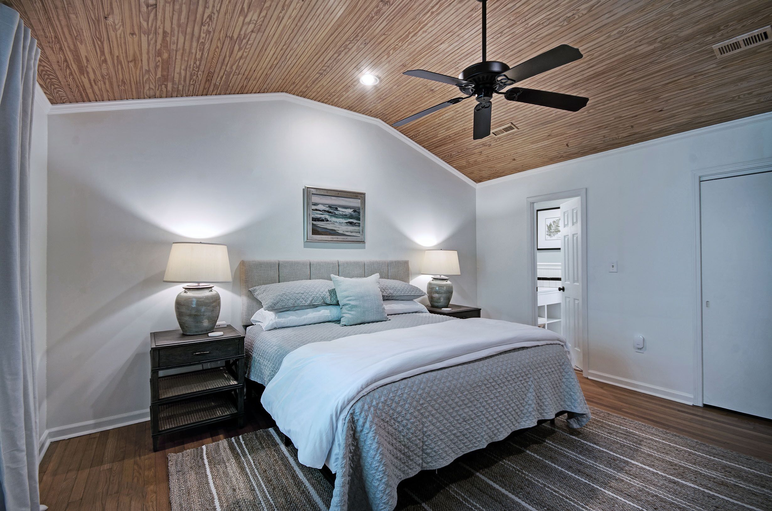 Une chambre avec un plafond en bois, un lit recouvert d’une couverture grise, deux tables de chevet avec des lampes et un tableau encadré au mur.
