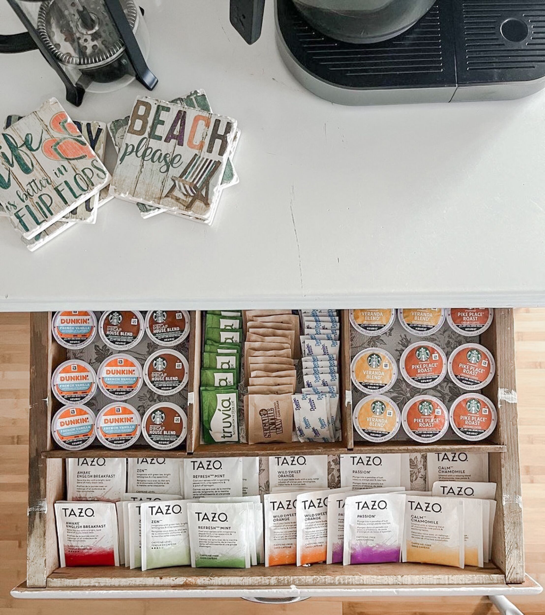 Uma bancada de cozinha com diversos produtos de café e chá, incluindo itens das marcas Dunkin' e Starbucks, e uma gaveta com vários sachês de chá Tazo.