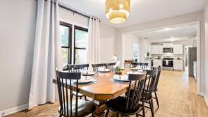 Une salle à manger avec une table en bois dressée pour six personnes, entourée de chaises. Un lustre est suspendu au plafond, et on aperçoit une cuisine au fond.