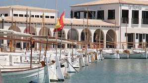 A marina with multiple boats docked, a building with arched windows, and a flag. - Holiday home Balaixa 8 by the sea (Puerto Pollensa)