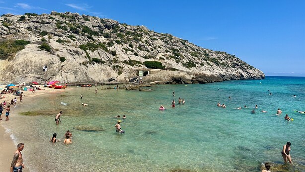 Uma praia com águas turquesa cristalinas, falésias rochosas e pessoas nadando e tomando sol.