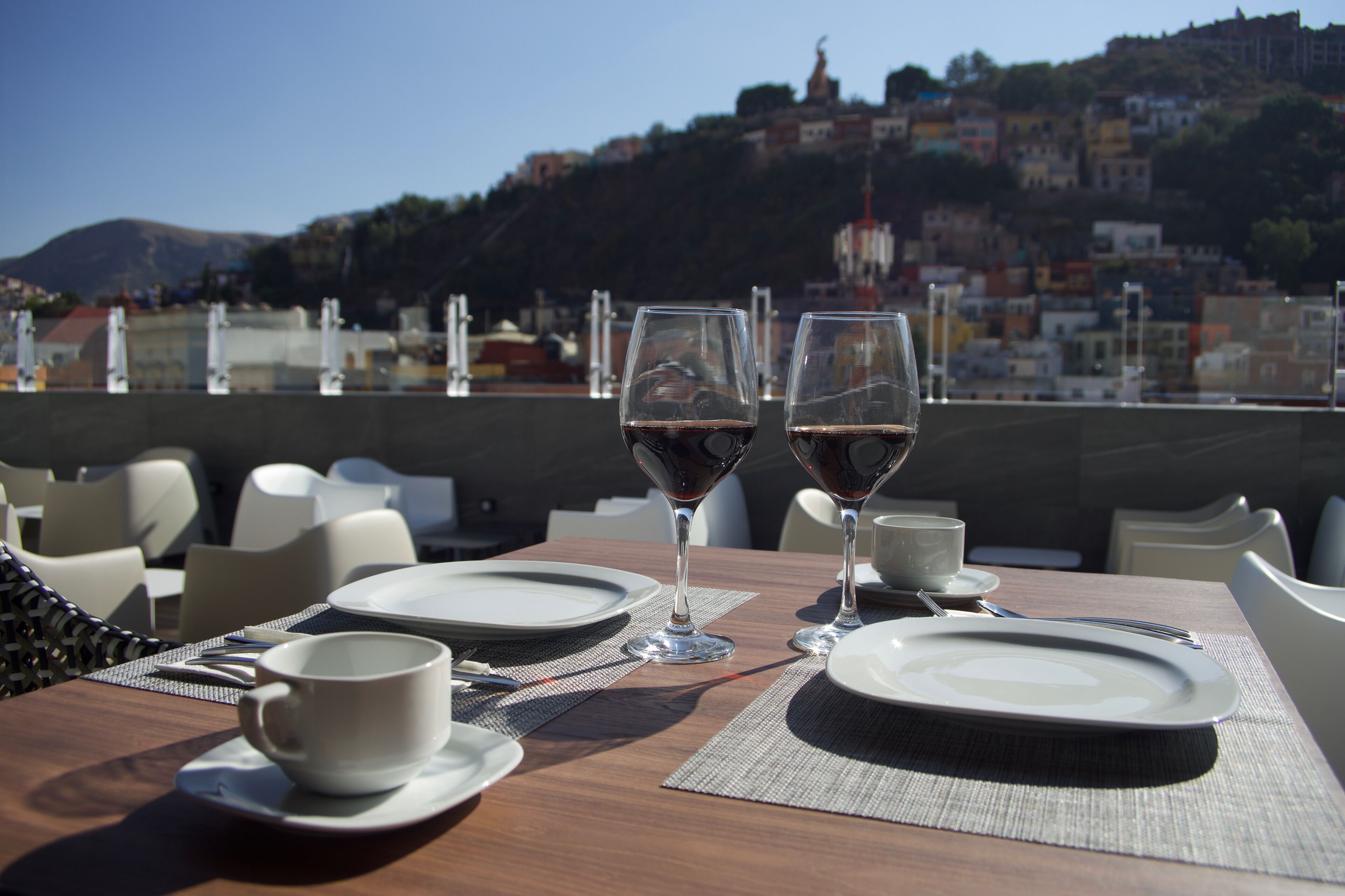 A dining table set with plates, glasses, and a cup, overlooking a cityscape with buildings and a hill.