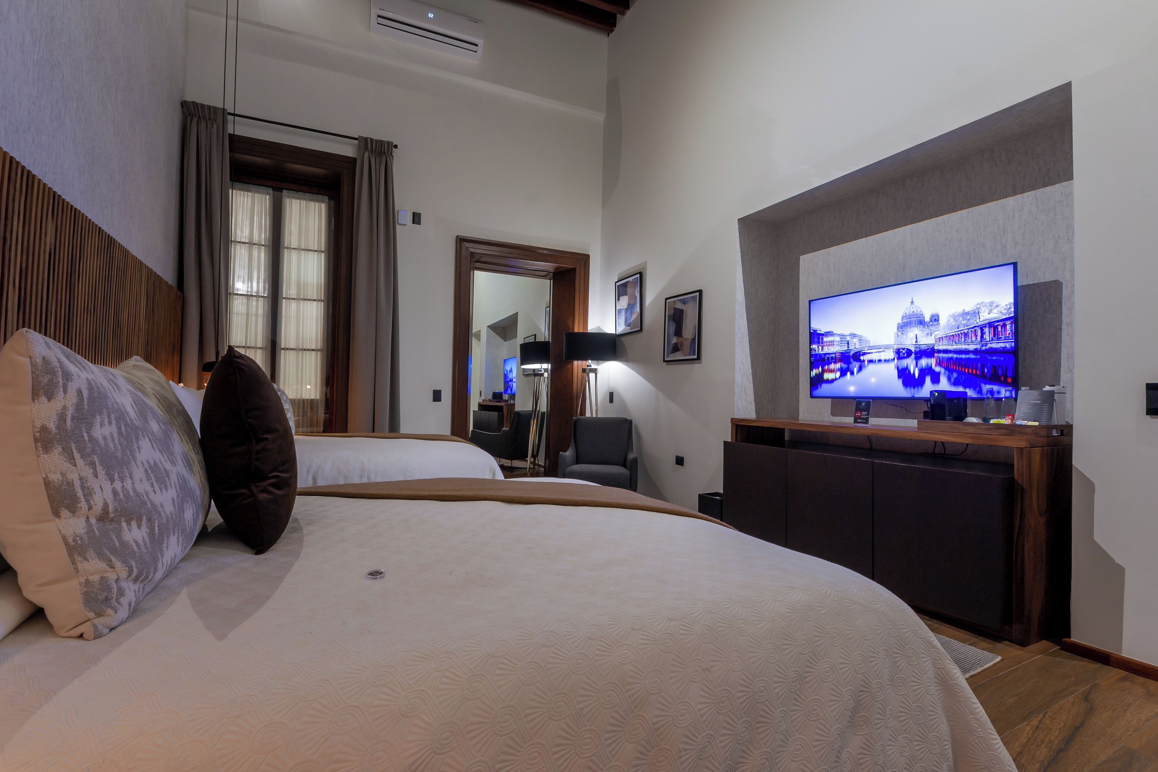 A hotel room with a large bed, a television mounted on the wall, a desk with a computer, and a window with curtains.