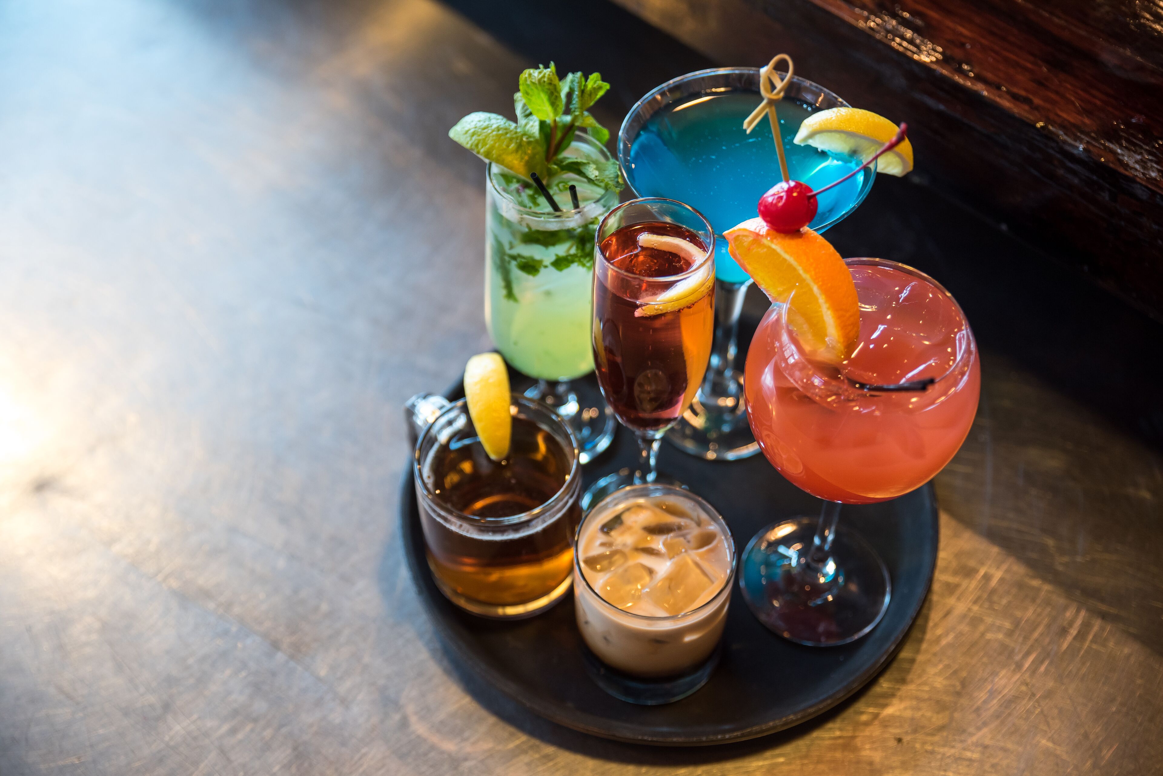 Variedad de bebidas sobre una bandeja, incluyendo un cóctel azul, una bebida rosada con adornito de naranja, y una bebida verde con menta.