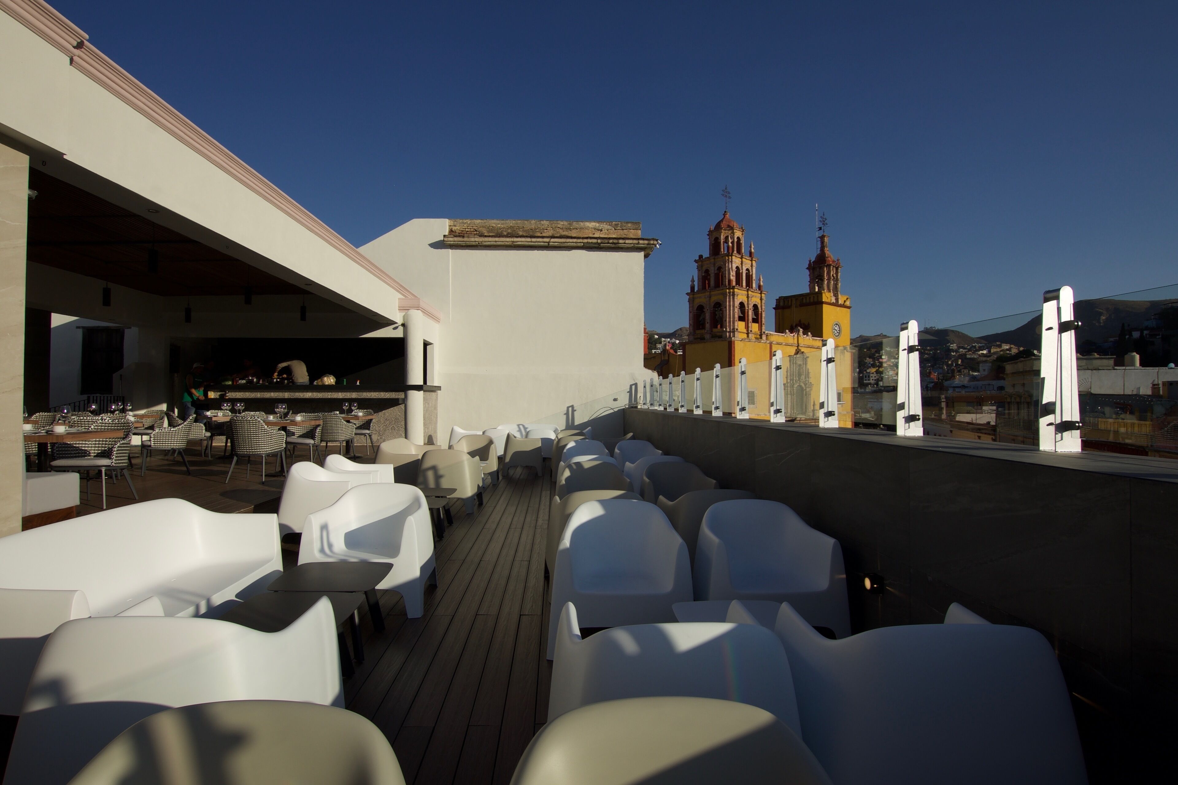 Terraza en la azotea con sillas y mesas blancas, un comedor y vistas a una iglesia con dos campanarios.