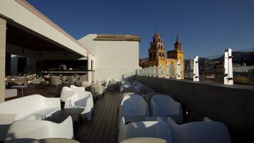 Terraza en la azotea con sillas y mesas blancas, un comedor y vistas a una iglesia con dos campanarios.