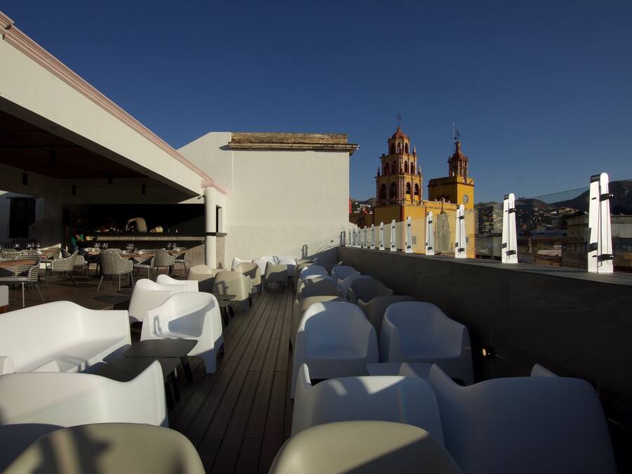 Terraza en la azotea con sillas y mesas blancas, un comedor y vistas a una iglesia con dos campanarios.