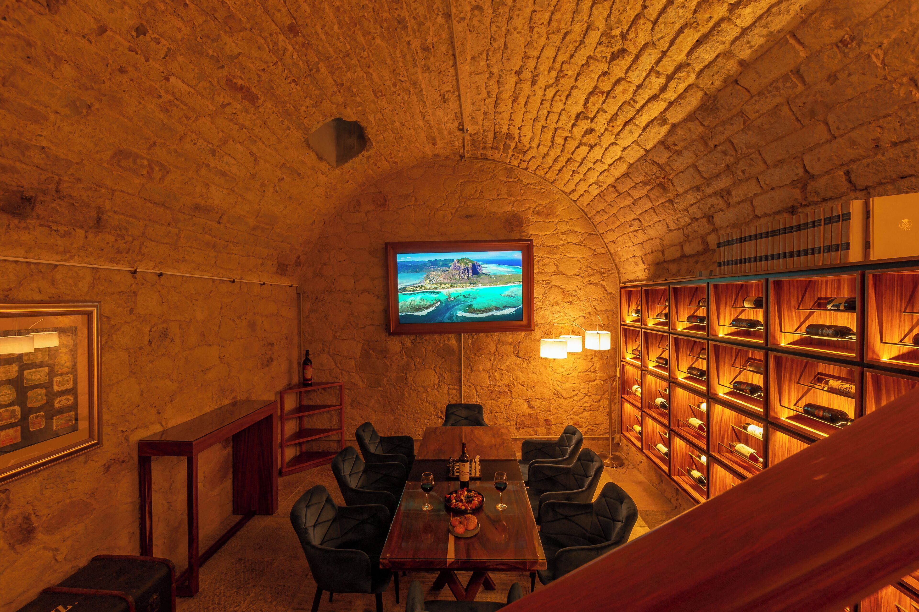 A stone-walled room with a wooden table and chairs, a television mounted on the wall, and a wine rack with bottles.
