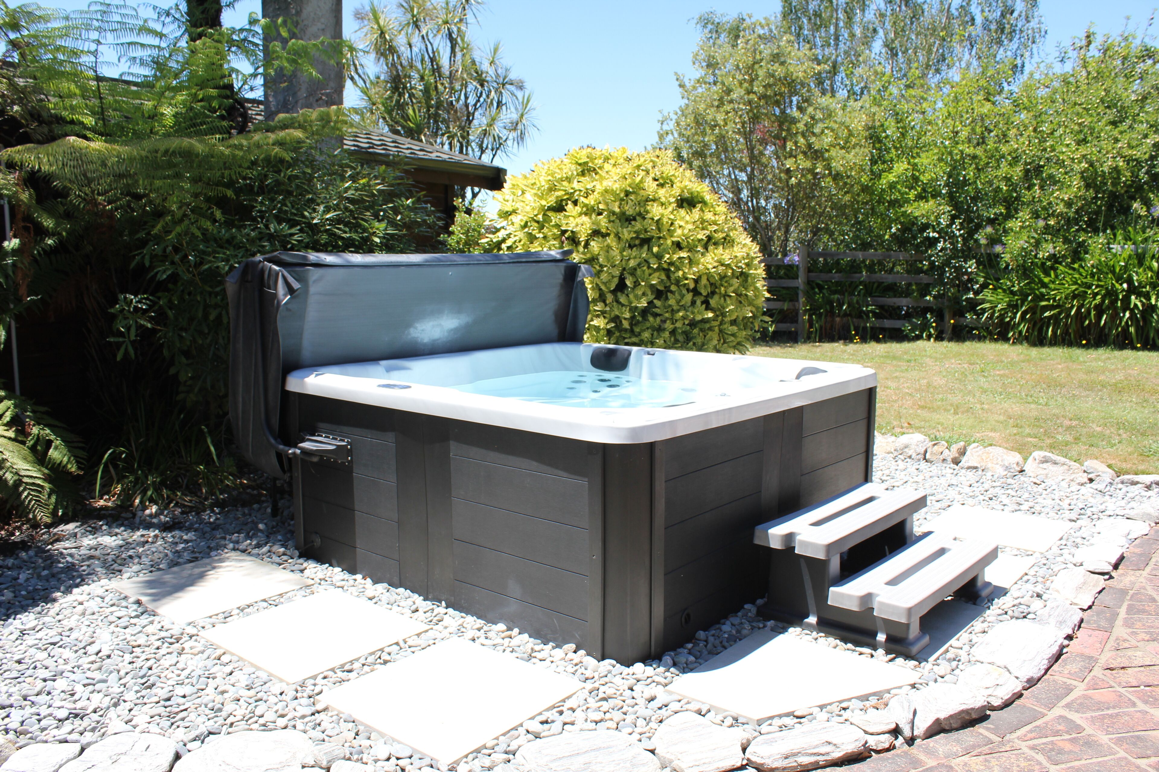 A hot tub in a garden setting with a stone pathway, surrounded by greenery and a wooden fence.