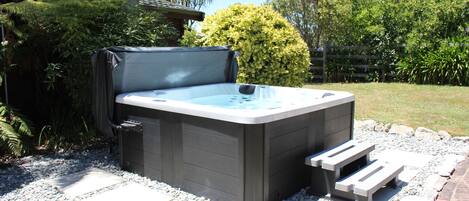 A hot tub in a garden setting with a stone pathway, surrounded by greenery and a wooden fence.