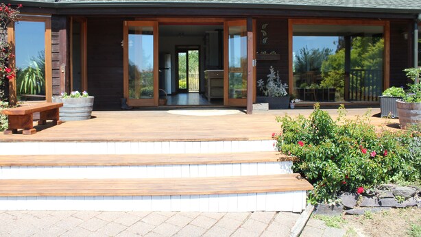 A modern house with a wooden deck, large sliding glass doors, and a patio area with steps leading to the entrance. - Large Country Home with Views - Spa Pool, Swimming Pool, Firepit, Games Room (Redwood Valley)