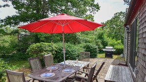 A wooden deck with a red umbrella, surrounded by wooden chairs and a table set for dining. - Spacious Home w/ Fire Pit and Central AC! (Eastham)