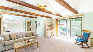 A spacious living room with a high ceiling, wooden beams, a sofa, a rocking chair, a glass coffee table, and a desk with a chair. - Spacious Home w/ Fire Pit and Central AC! (Eastham)