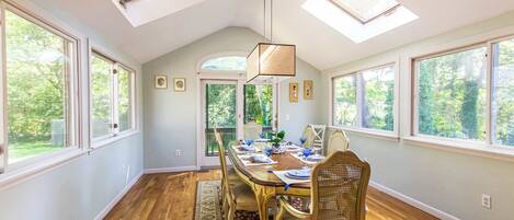 A dining room with a large table, chairs, and a chandelier. There are skylights and large windows providing natural light.