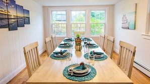 A dining room with a long wooden table set for six, flanked by wooden chairs. The room has white walls, large windows with a view of greenery, and artwork on the walls. - 100 Ft to Skaket Beach! (Orleans)