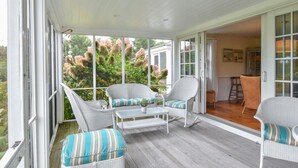 Un porche cubierto con muebles de mimbre blanco, una mesa de centro de vidrio y vistas a un jardín con flores rosadas.
