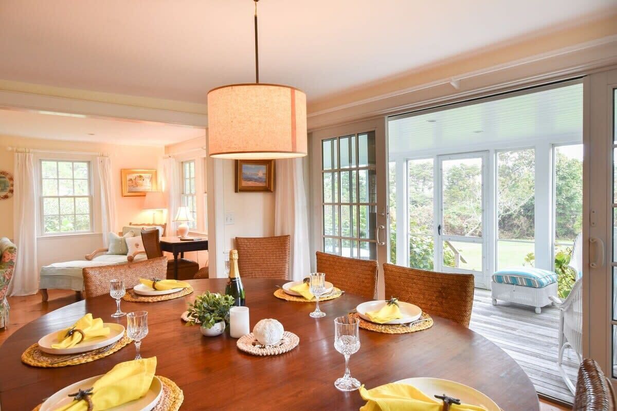 A dining room with a wooden table set for six, surrounded by wicker chairs, with a large window offering a view of the outdoors.