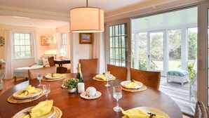 Comedor con mesa de madera dispuesta para seis comensales, rodeada de sillas de mimbre y un ventanal con vistas al exterior.