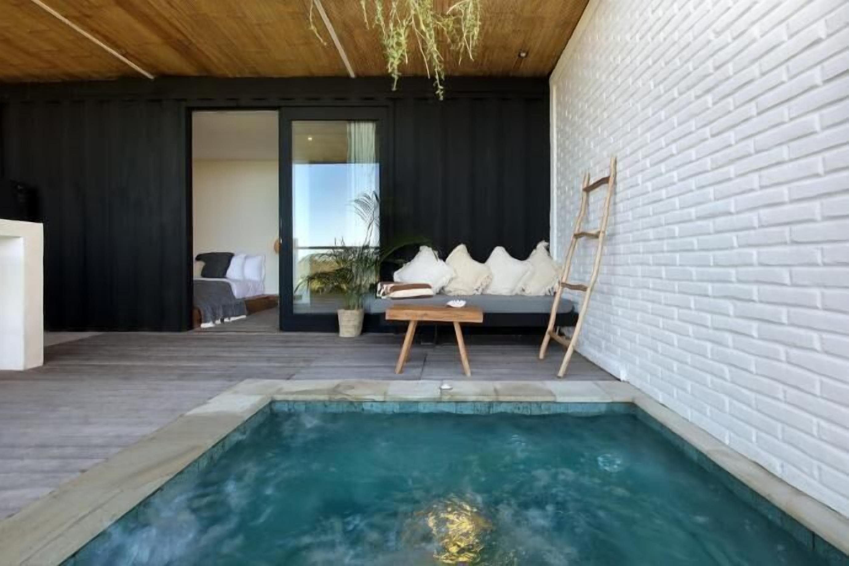 A modern interior with a pool, a sofa with cushions, a wooden table, and a white brick wall.