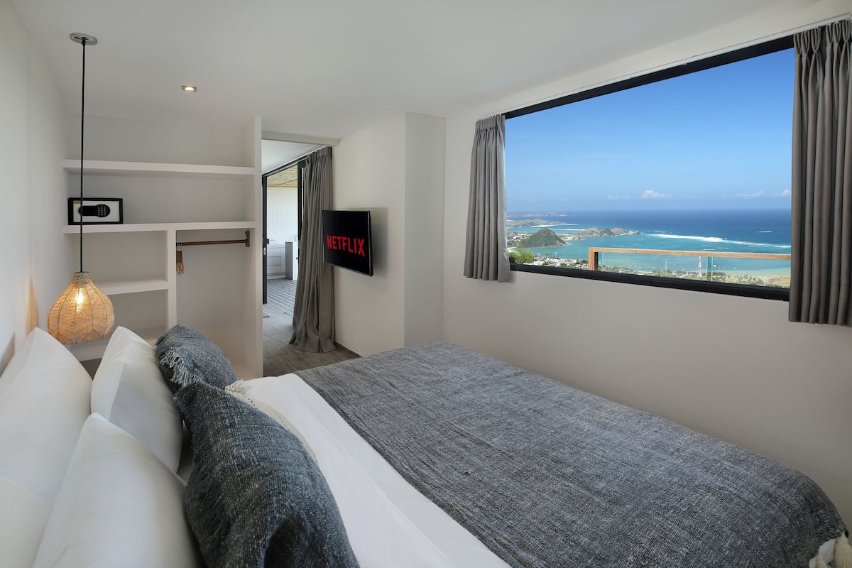 Un dormitorio con una ventana grande, una cama, un televisor y vista al océano.