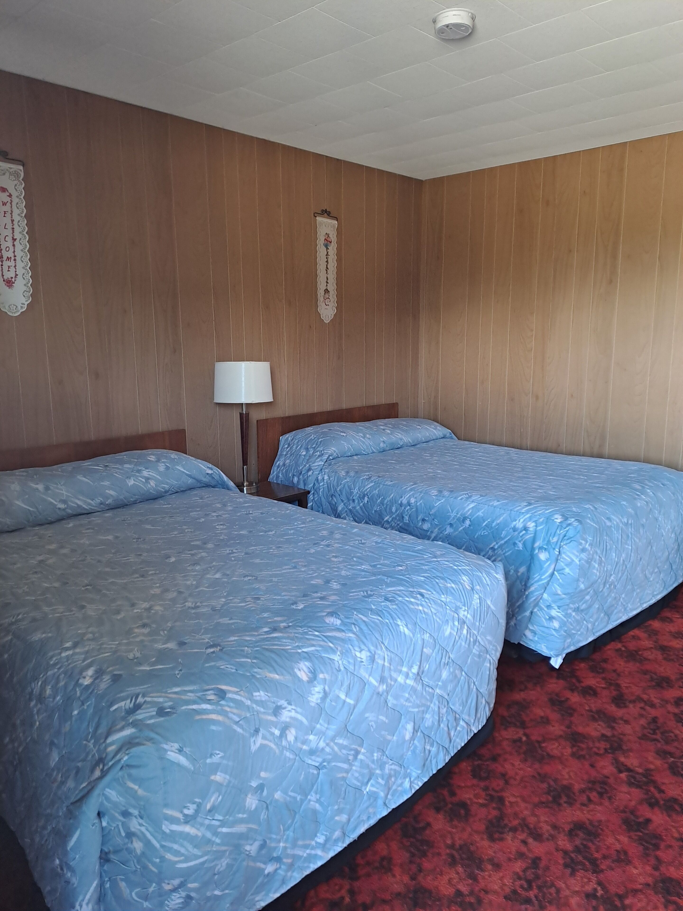 A hotel room with two beds, a red carpet, wooden panel walls, a white ceiling, and a wall-mounted lamp.