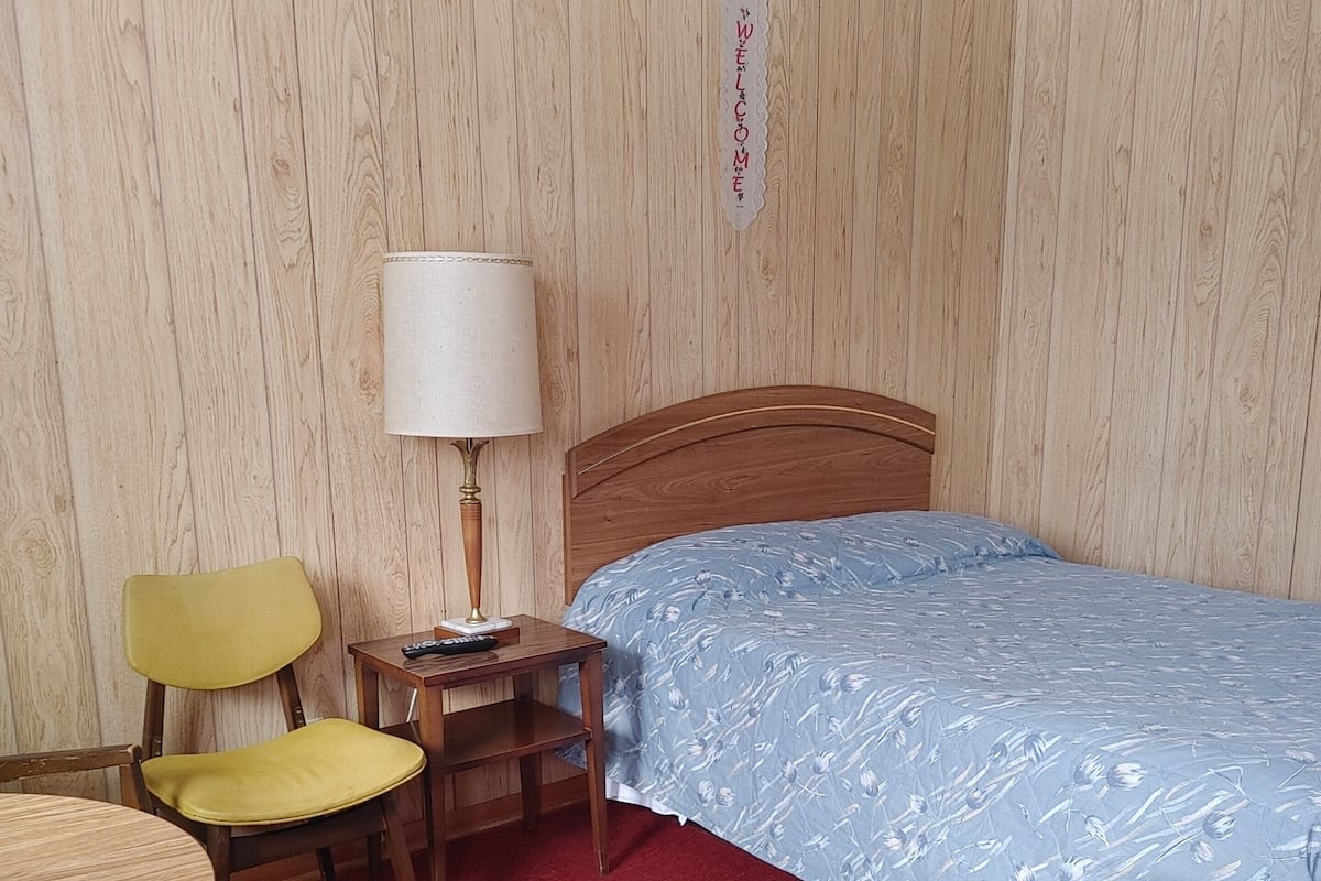 Habitación de hotel con cabecera de madera, cama con colcha floral azul, una silla amarilla, una mesita de madera con lámpara y alfombra roja.