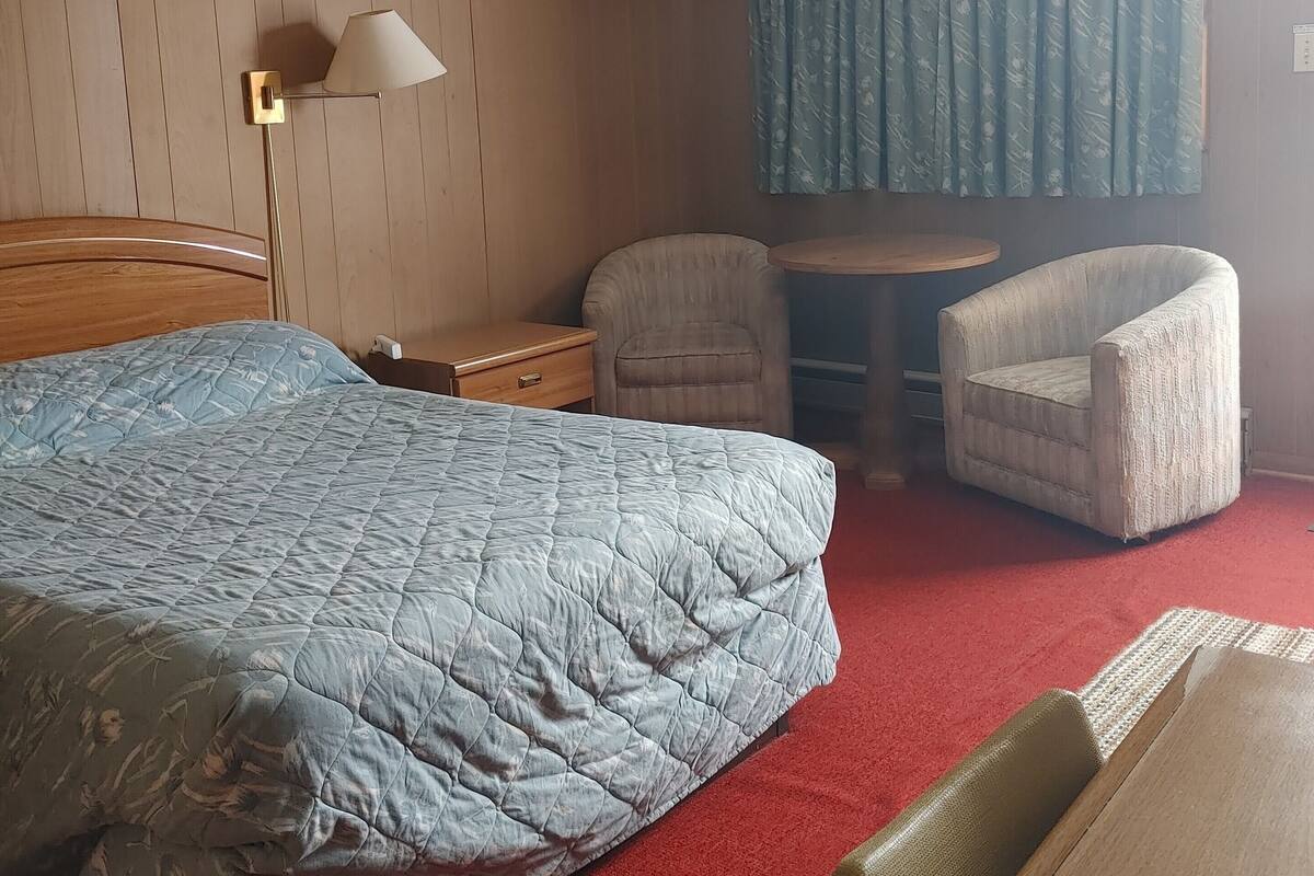 Una habitación de hotel con cama, escritorio, silla, mesita, lámpara y ventana con cortinas.