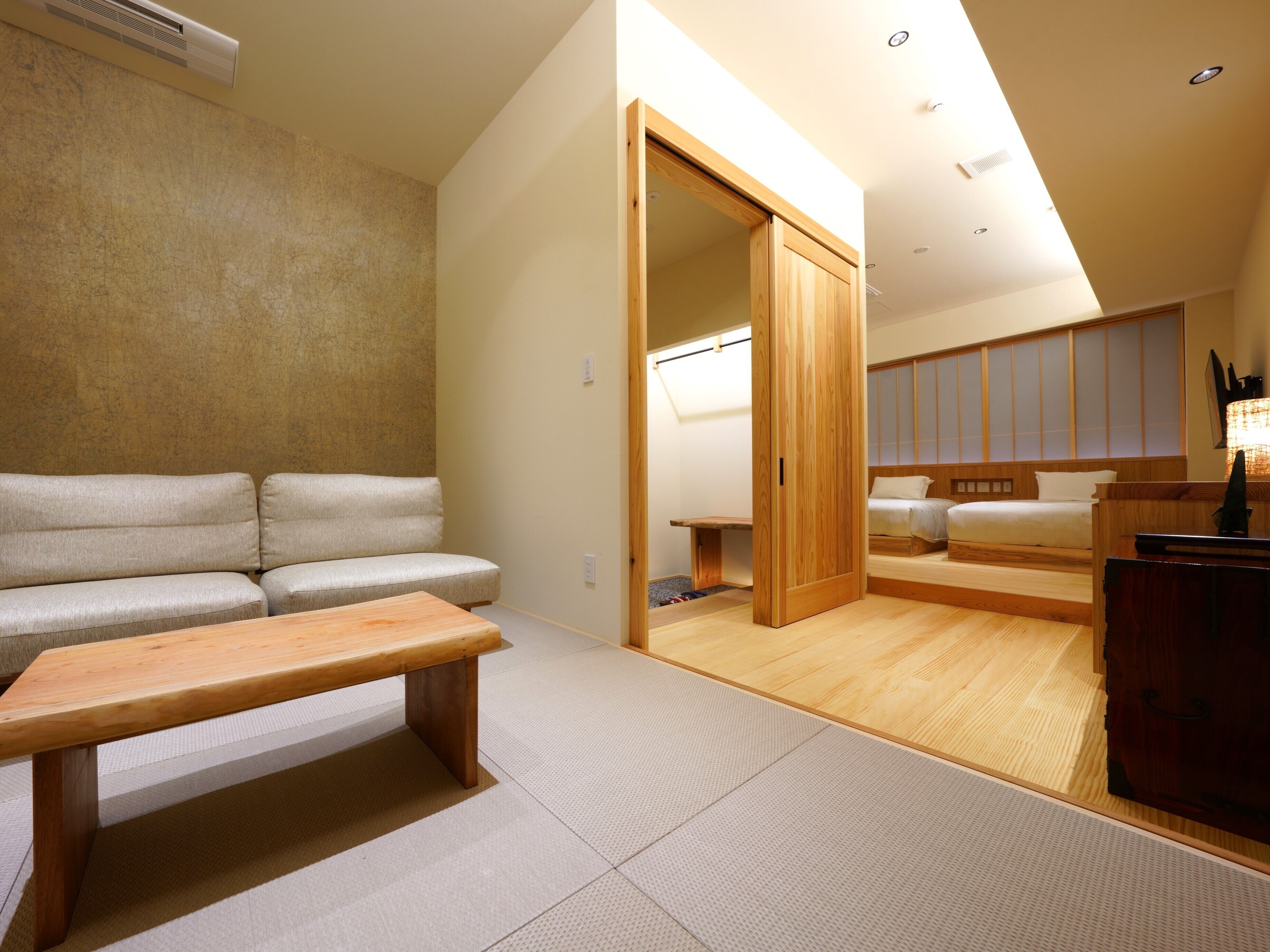 Een moderne hotelkamer met een bank, een houten zitbank, een bed en een schuifdeur naar een badkamer.