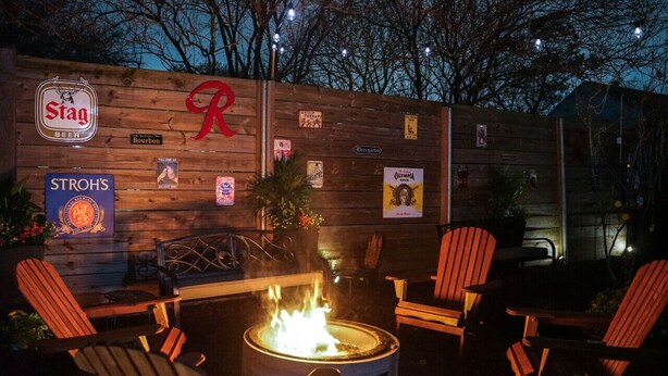 Un área de estar exterior de madera con sillas Adirondack, un hogar para fuego y varios carteles y anuncios en la pared.
