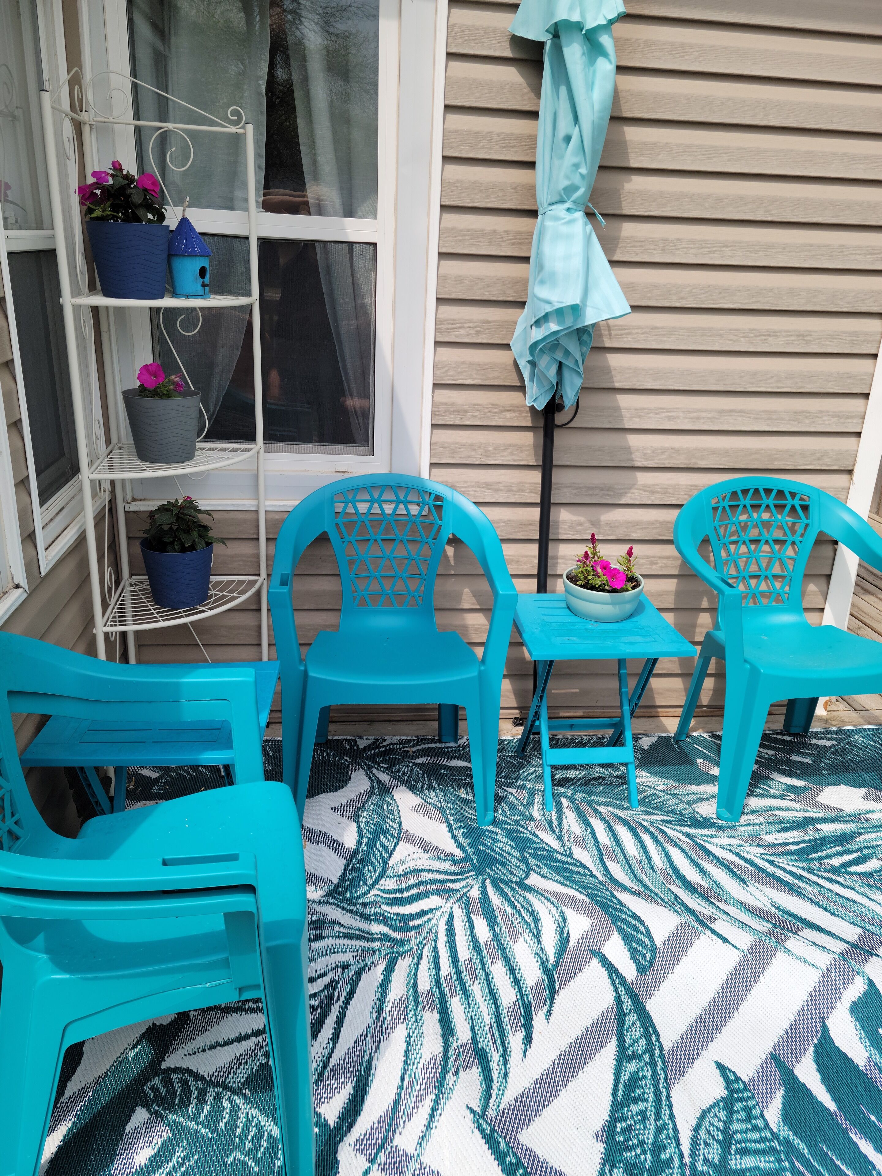 Ein kleiner Patio-Bereich mit türkisfarbenen Stühlen und einem Tisch, ein weißes Metallregal mit Topfpflanzen und ein Fenster mit Vorhang.