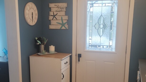 Un pasillo con suelo de madera, una puerta blanca con ventanal de vidrio, un reloj de pared y un cartel de bienvenida.