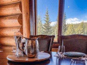 Un intérieur en bois avec une table dressée pour le repas, une carafe et une vue sur une forêt à travers de grandes fenêtres.