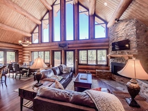 Un intérieur spacieux de chalet en bois, doté d’une cheminée, de canapés en cuir, d’un coin repas et de grandes fenêtres.