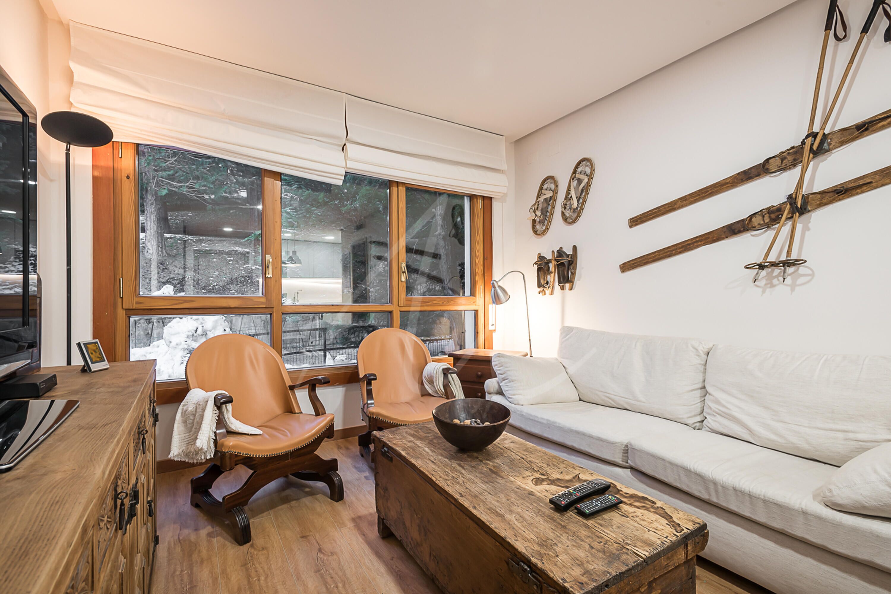 Een gezellige woonkamer met een houten salontafel, twee leren fauteuils, een witte bank en een groot raam met uitzicht op bomen.