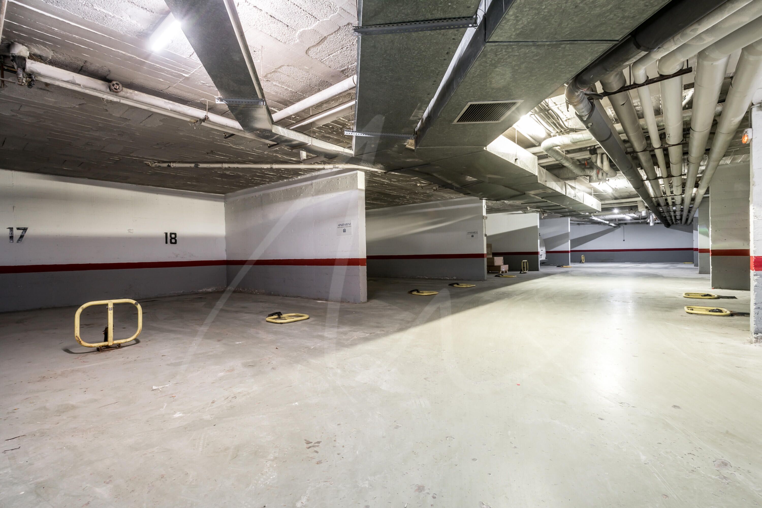Un aparcamiento de varios niveles con suelos y paredes de hormigón, plazas de aparcamiento numeradas y elementos estructurales visibles como vigas y tuberías.