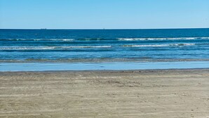 Una playa con olas, cielo despejado y barcos a lo lejos.