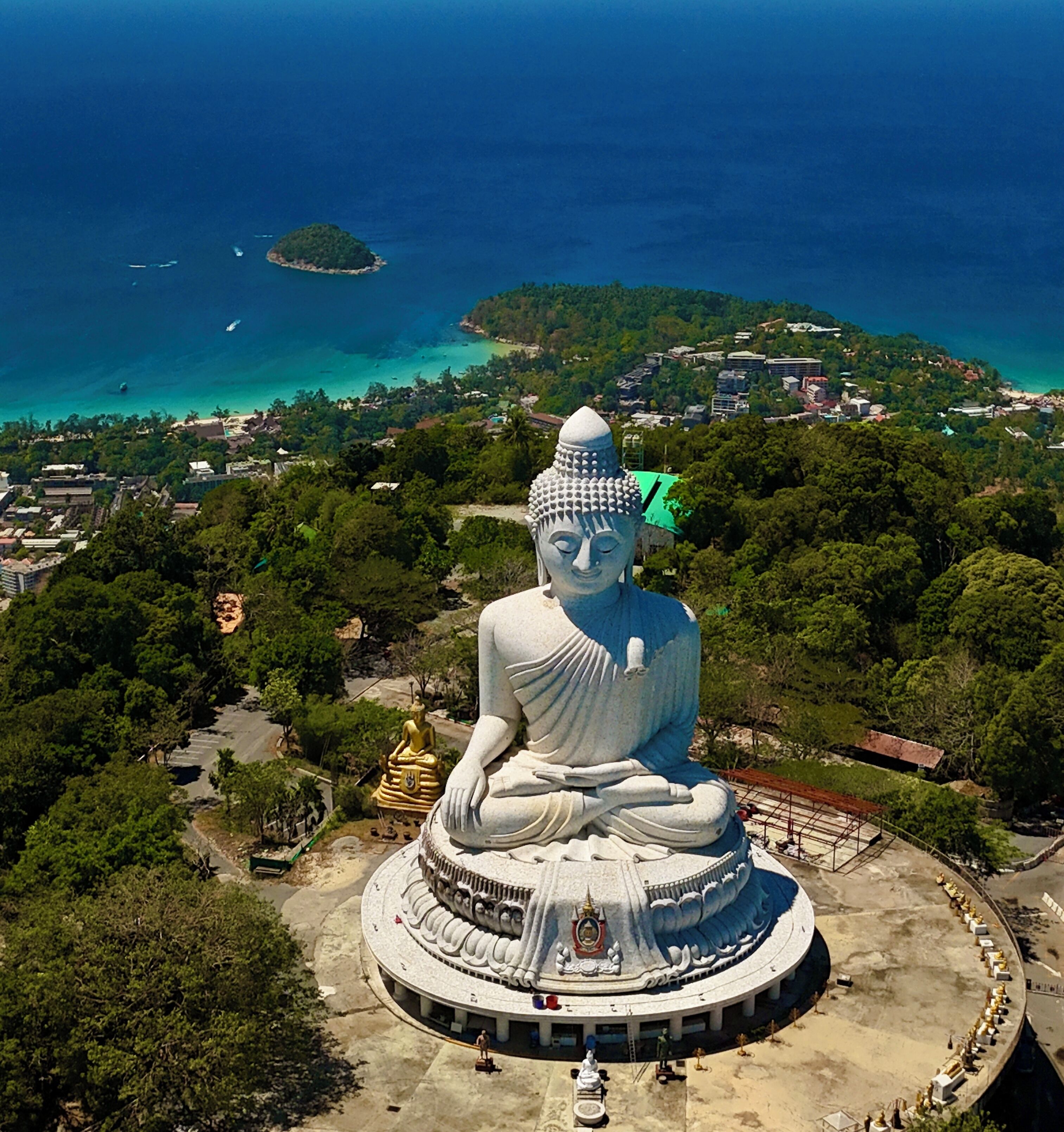 En stor, vit Buddha-staty omgiven av en rundpromenad, med utsikt över en kuststad och turkosblått vatten i bakgrunden.