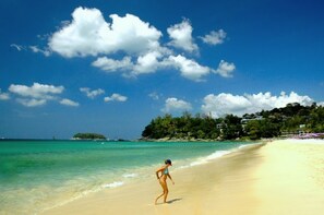 Een strand met helder turquoise water, een vrouw in een bikini en een kustlijn met huizen.